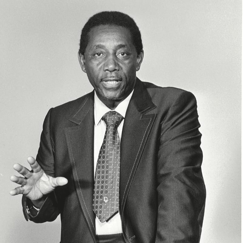 A man in a suit and tie gestures with his left hand in front of a plain background.