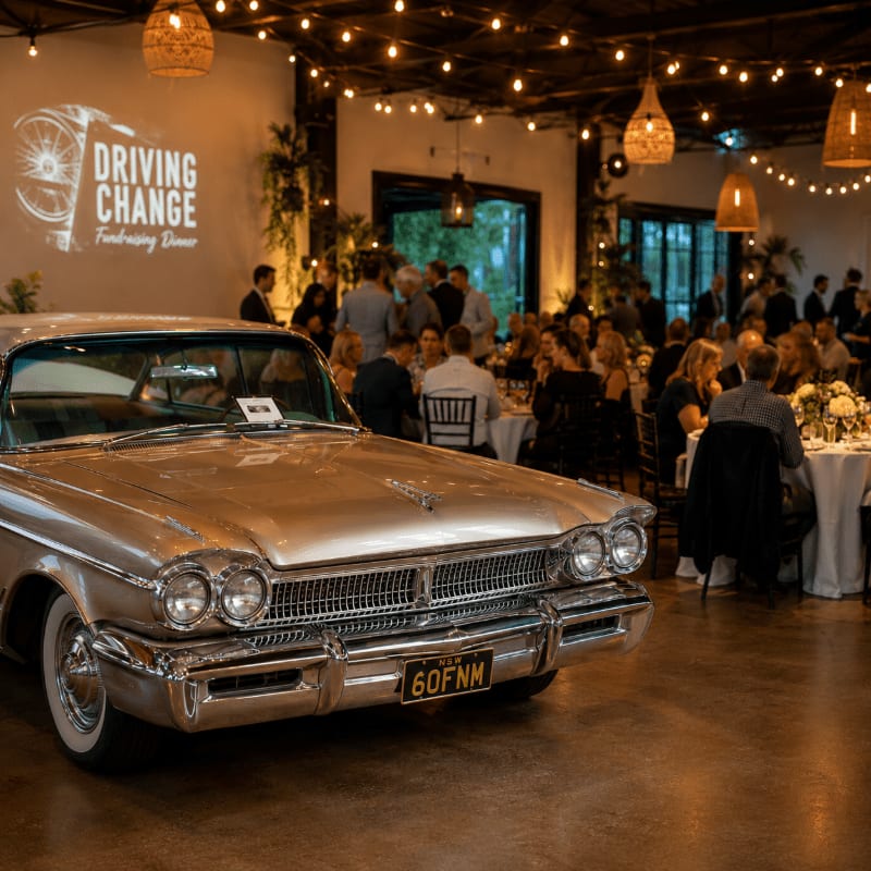 A vintage car is parked in the foreground of a dimly lit ballroom where a fundraising dinner is in progress, with guests seated at round tables and a "Driving Change" logo projected on the back wall.
