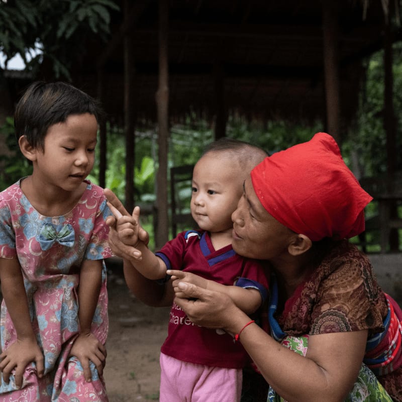 A woman in a red headscarf gently holds a baby while an older child looks on, with a thatched-roof structure and lush greenery in the background.