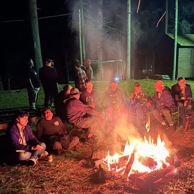 A group of people sit around a crackling campfire in a clearing at night, with trees and a small building visible in the dark background.