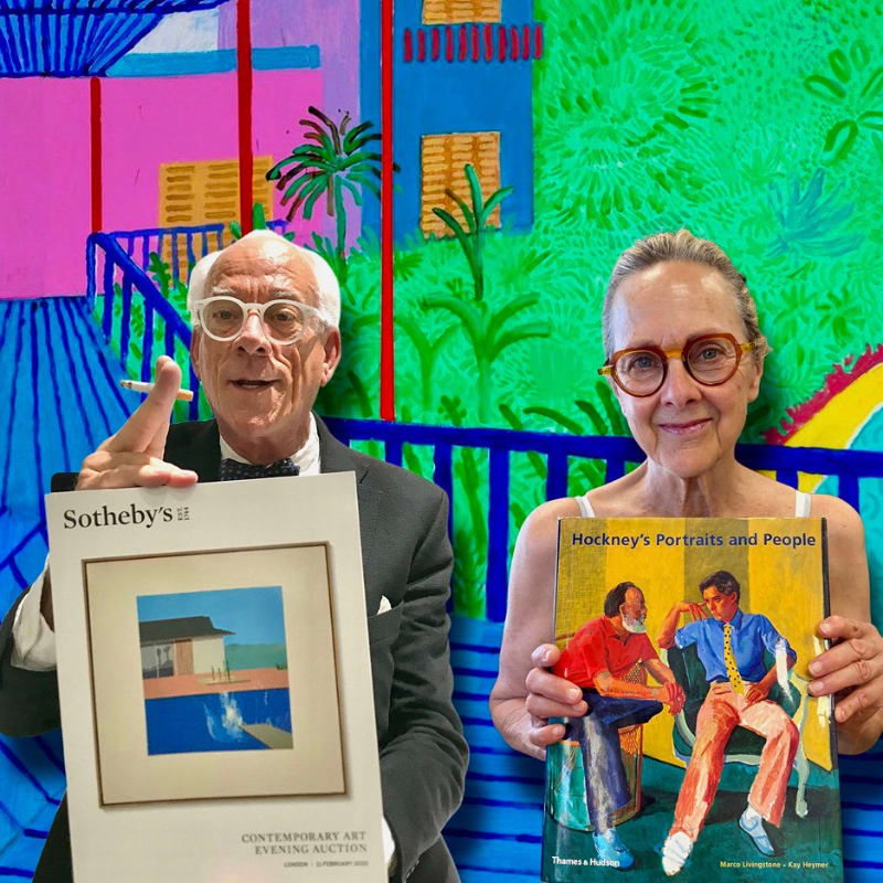 An older man in a suit and an older woman in a tank top hold up a Sotheby's auction catalog and a book of David Hockney's portraits, respectively, in front of a vibrant, colorful mural.