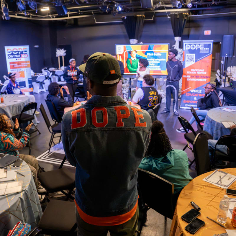 In the foreground, a person wearing a denim jacket with "DOPE" on the back faces away from the camera, while the background shows an audience seated at tables watching a presentation on a large screen.