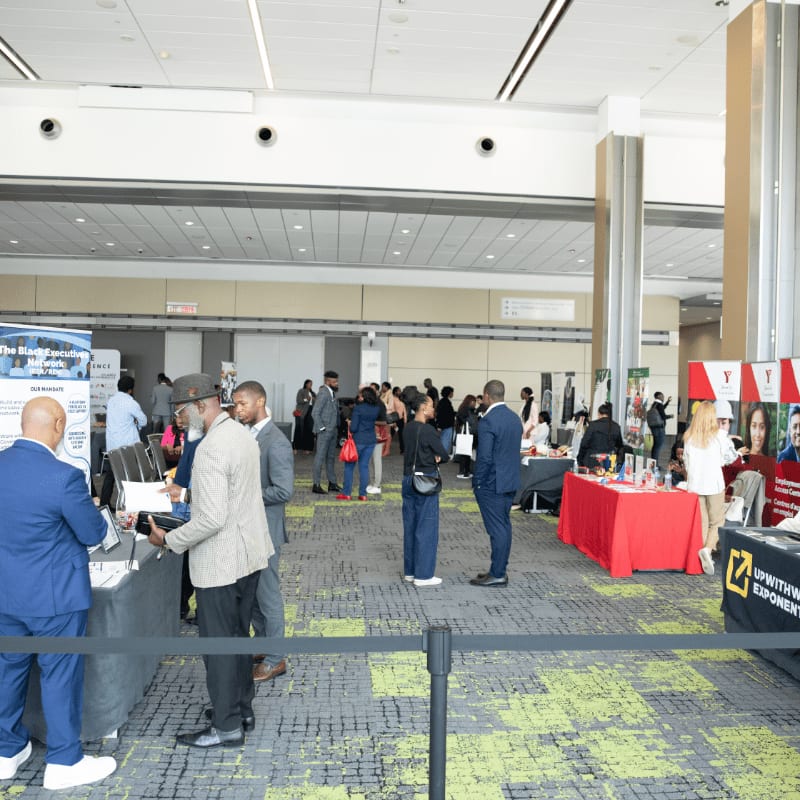People in business attire converse at booths in a large convention hall with a green and black patterned carpet.