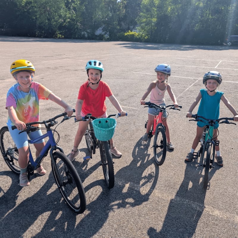 Four children in colorful helmets and outfits pose with their bicycles on a paved surface, with trees and a building visible in the sunny background.