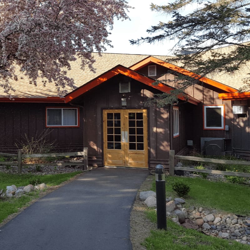 A paved walkway leads to a brown building with double wooden doors, framed by a wooden fence and blooming trees in the foreground, while the background features more of the building and surrounding trees.