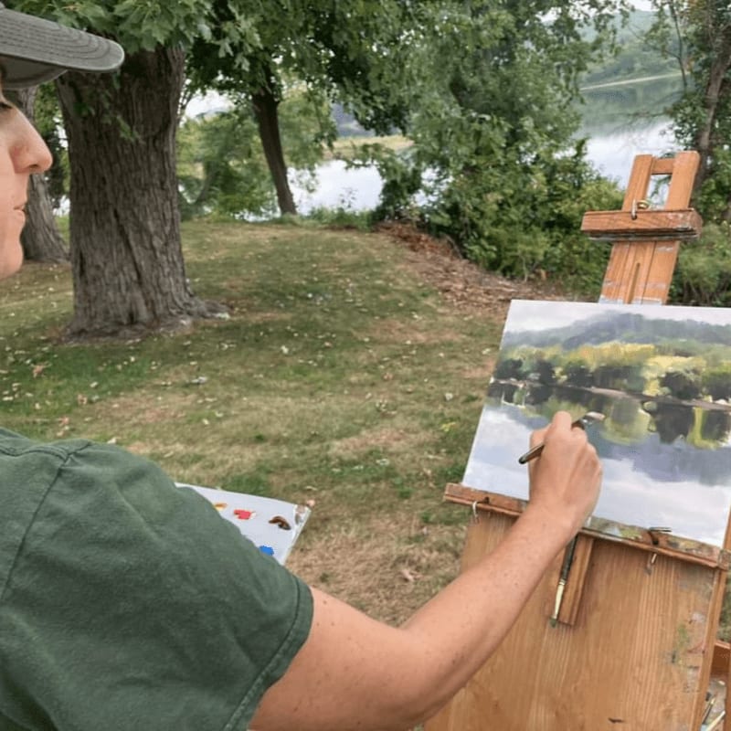 A person in a green shirt and gray hat paints a landscape of trees and water on an easel in a grassy area with a lake in the background.