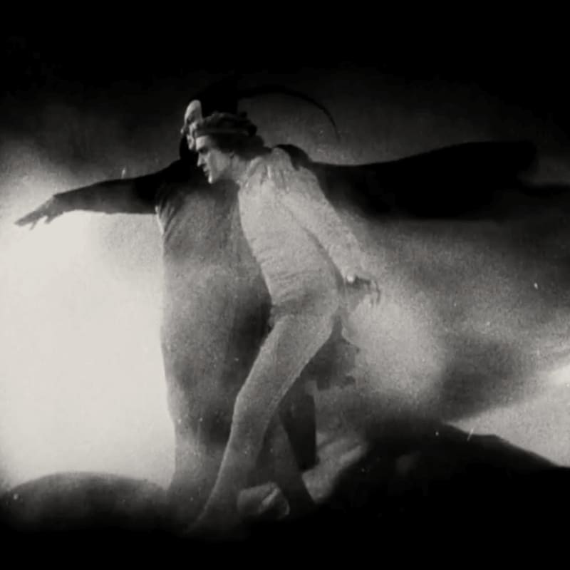 A man in a light-colored suit is embraced by a shadowy figure with outstretched arms against a dark, smoky background.