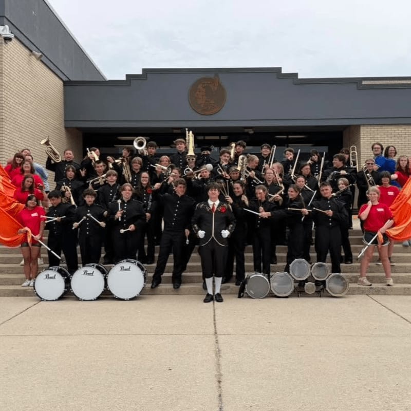 A marching band with instruments and flags stands on steps in front of a school building.