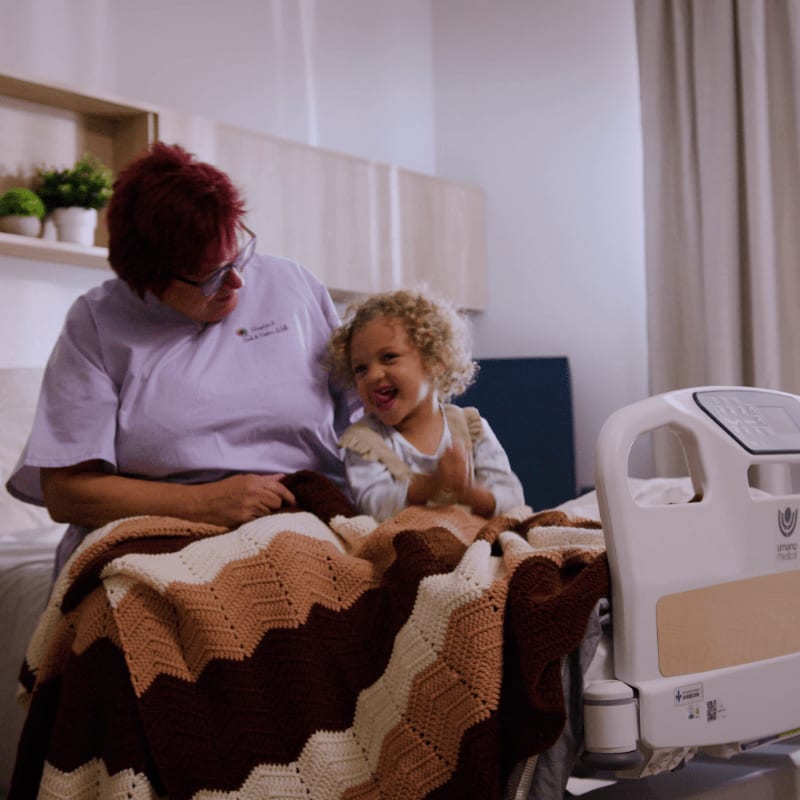 A woman with red hair and glasses sits beside a laughing child on a hospital bed covered with a chevron-patterned blanket, with shelves and a curtain visible in the background.
