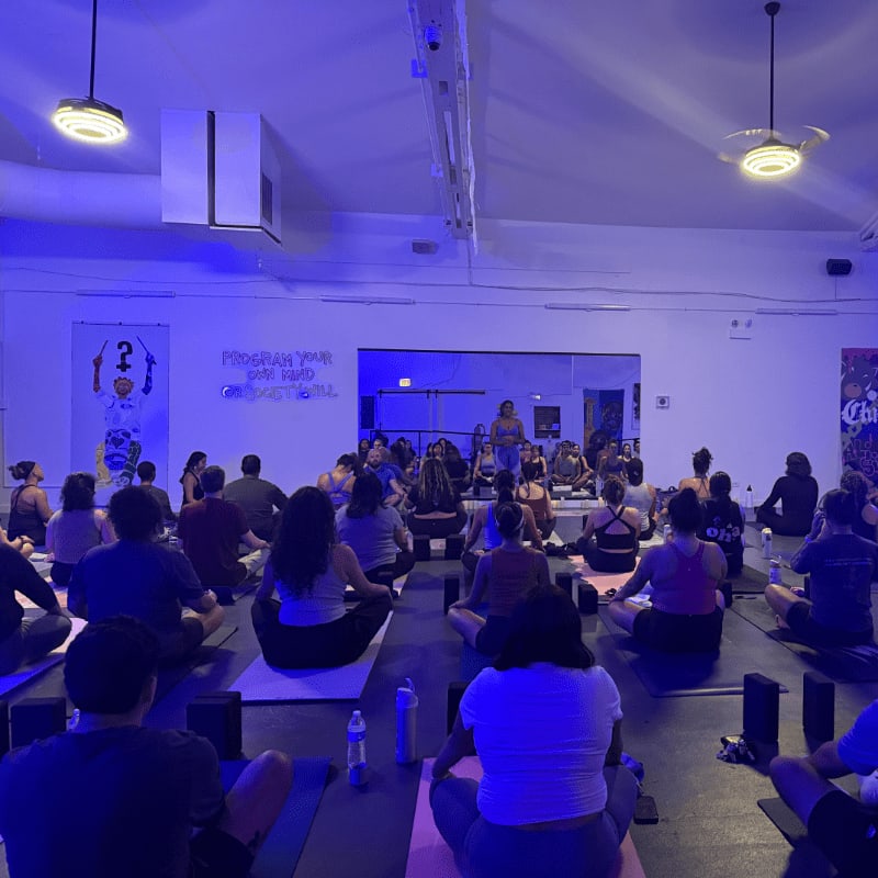 In the foreground, people sit cross-legged on yoga mats in a dimly lit room, while in the background, a woman stands addressing the group in front of a mirrored wall.