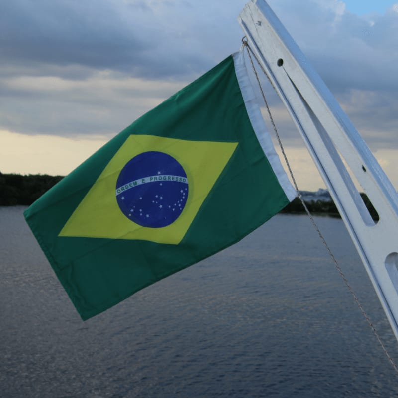 A Brazilian flag flutters in the foreground against a backdrop of calm water and a cloudy sky.