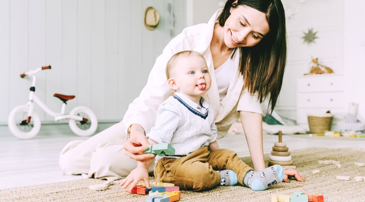 A mother and her baby play with colorful blocks on a rug in a bright room with a toy bicycle in the background.