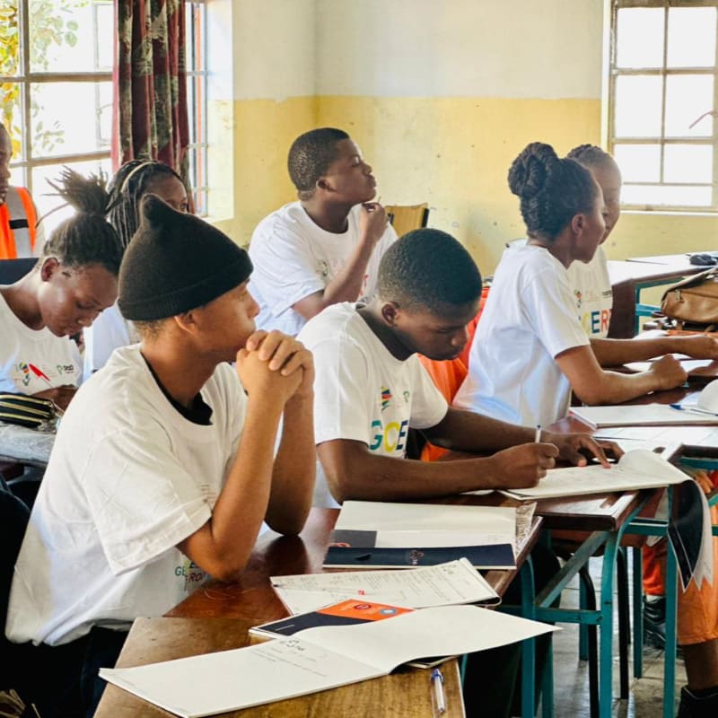 Students in white t-shirts are seated at desks in a classroom, with windows and a whiteboard visible in the background.