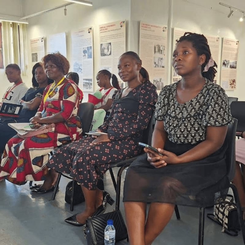 Several women are seated in chairs in the foreground, facing a wall with framed exhibits in the background.