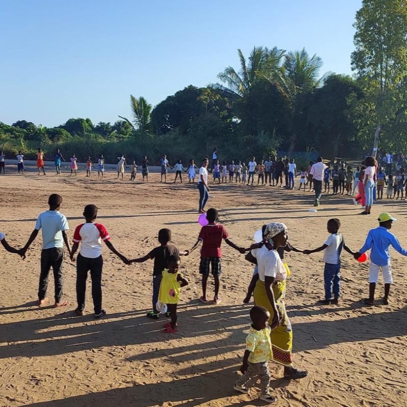 Children and adults hold hands in a circle on a sandy ground in the foreground, with a line of people and trees in the background under a clear blue sky.
