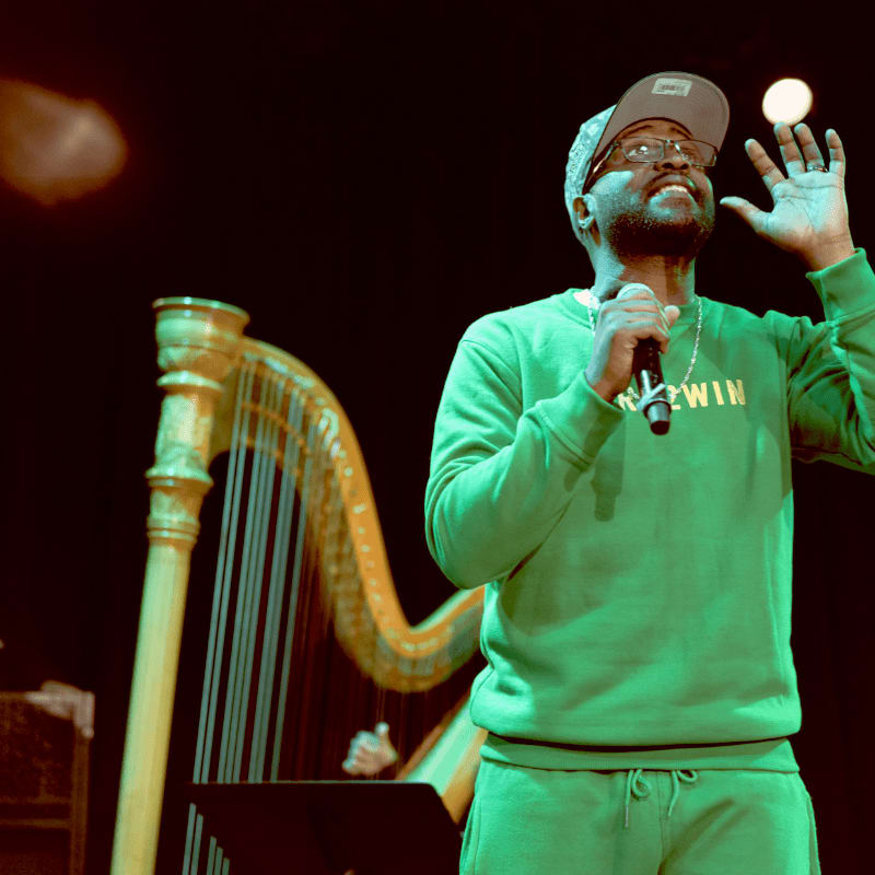 A man in a green sweatshirt and cap holds a microphone and a small white object in his raised hand, with a golden harp visible in the blurred background.