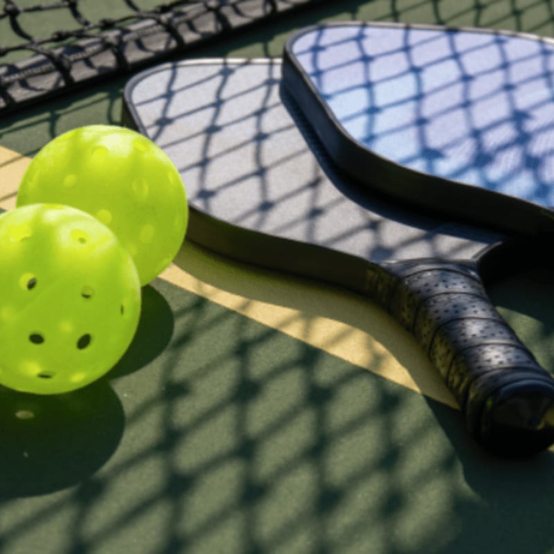 Two bright green pickleballs rest on a court next to two paddles, with a net casting a shadow over the scene.