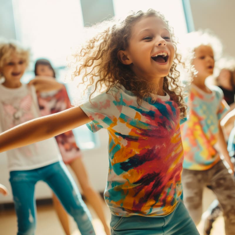 A joyful young girl with curly hair in a tie-dye shirt dances in the foreground, while other children in colorful attire move around her in a bright, sunlit room.