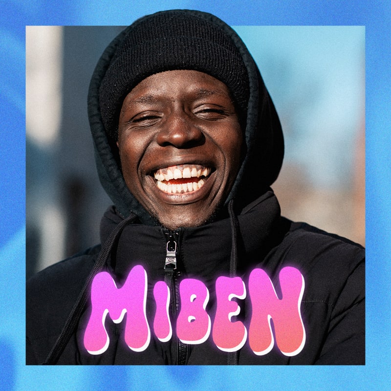 A man in a black beanie and jacket laughs heartily in the foreground, with a blurred urban background.