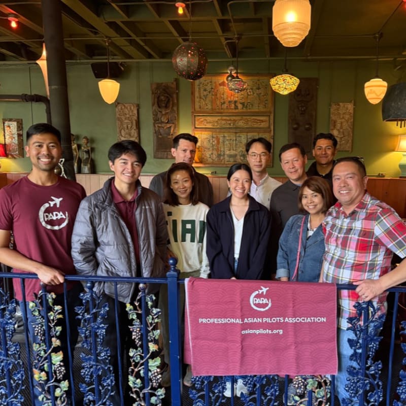 A group of ten people stand behind a decorative railing in the foreground, with a banner displaying "PROFESSIONAL ASIAN PILOTS ASSOCIATION" in front of them, while ornate decorations and lighting adorn the green-walled background.