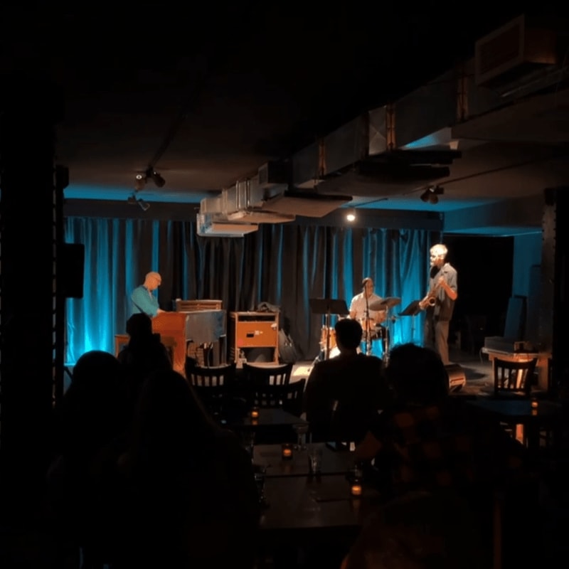 Musicians perform on a stage with blue lighting and curtains in the background, while an audience sits in the dimly lit foreground.