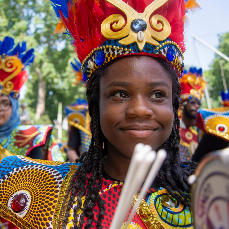 A young person in a vibrant headdress and costume smiles as they hold drumsticks, with other costumed individuals and trees visible in the blurred background.