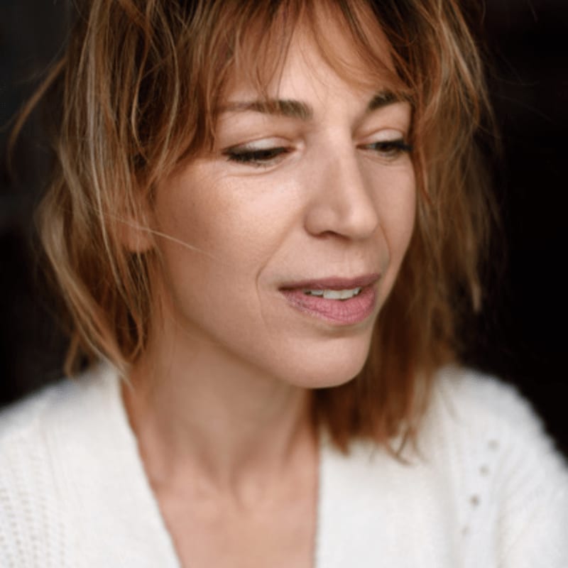 A woman with short, tousled brown hair and a white sweater is looking down against a dark background.