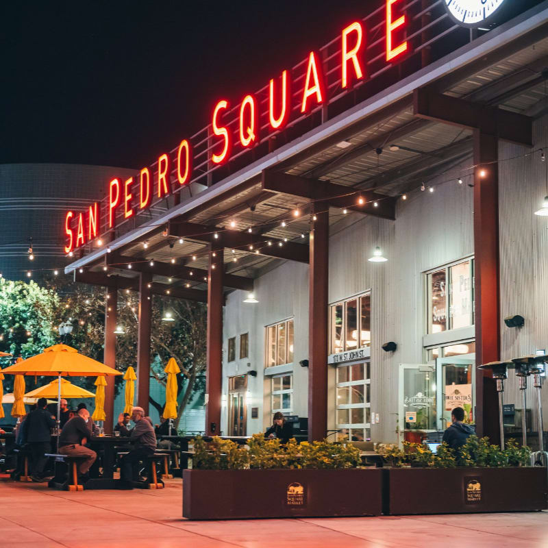 People dine under yellow umbrellas in the foreground, while the "SAN PEDRO SQUARE" sign glows in neon above a modern building in the background.