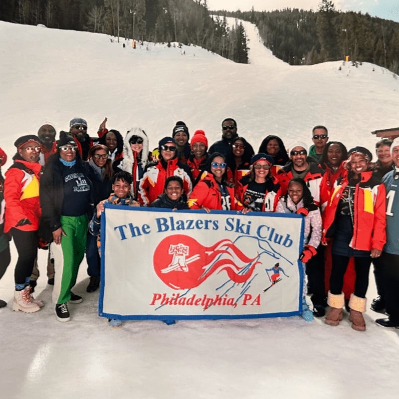 A large group of people in ski attire pose for a photo in front of a ski slope, holding a banner that reads "The Blazers Ski Club Philadelphia, PA."