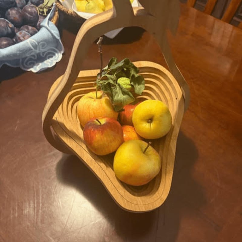 A wooden fruit basket filled with apples sits on a wooden table, with a basket of plums visible in the background.