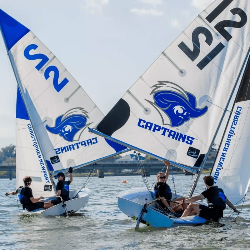 Two sailboats with blue and white sails bearing the "Captains" logo race on a lake with a bridge in the background.