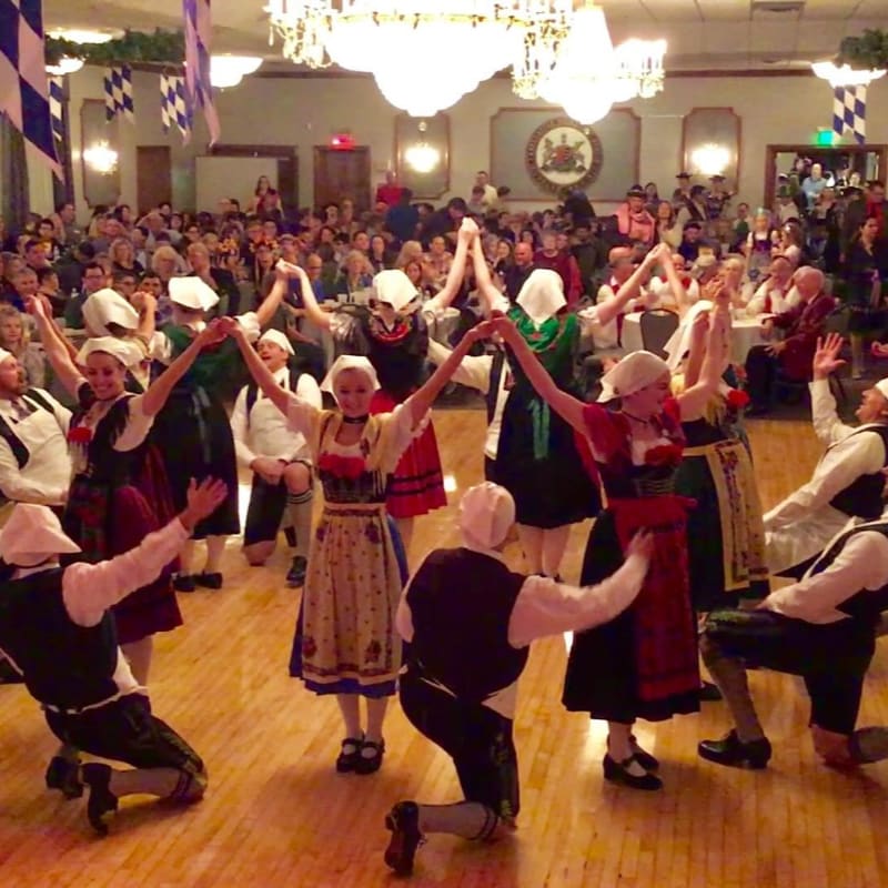 A group of people in traditional German attire dance in a circle on a wooden floor, while a larger crowd watches in the background of a decorated hall.