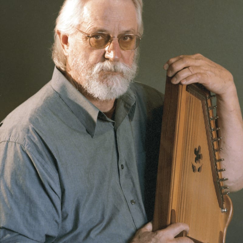 A bearded man with glasses holds a zither against a plain green background.