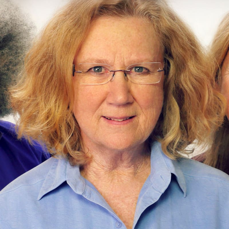 A woman with glasses and a blue shirt smiles in the foreground, with the blurred shoulders of two other people visible in the background.
