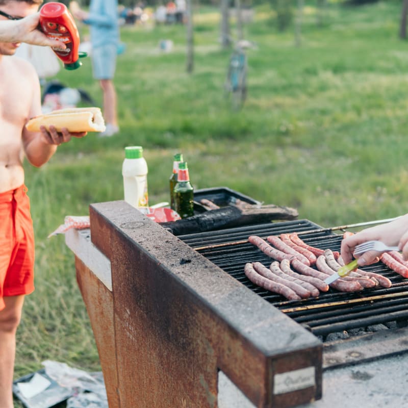 man wearing red shorts standing at grill putting ketchup on hotdog