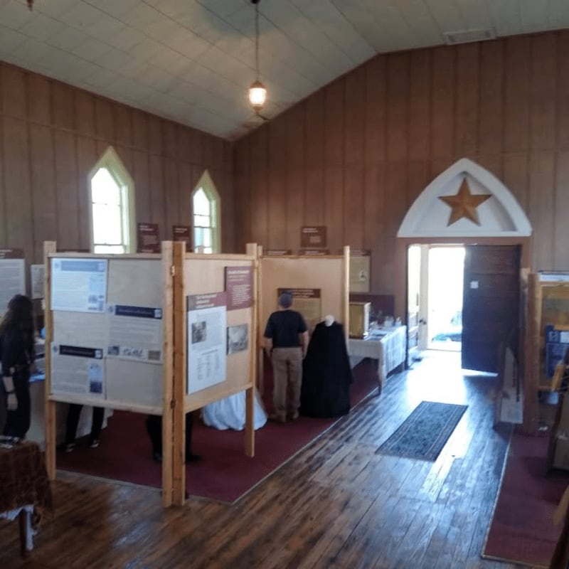 People are viewing exhibits in a wooden-paneled room with arched windows and an open doorway leading outside.