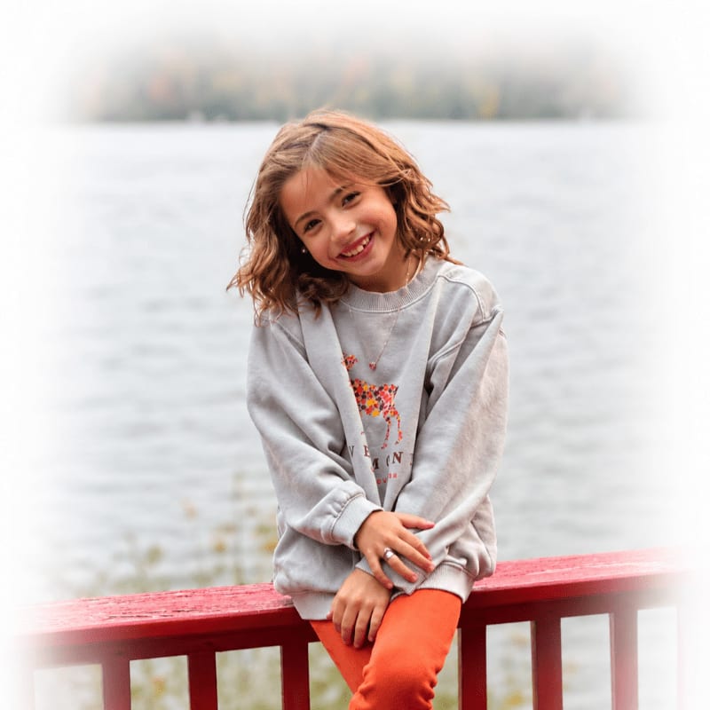 A smiling young girl in an orange top and grey sweatshirt sits on a red railing with a blurred lake and trees in the background.