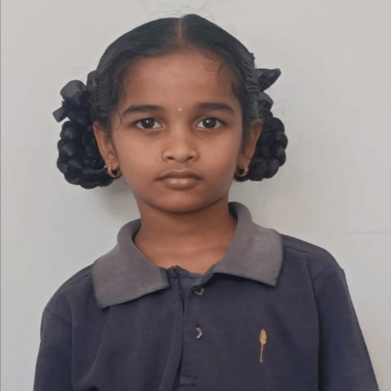 A young girl with braided pigtails and a dark collared shirt stands against a plain, light-colored background.