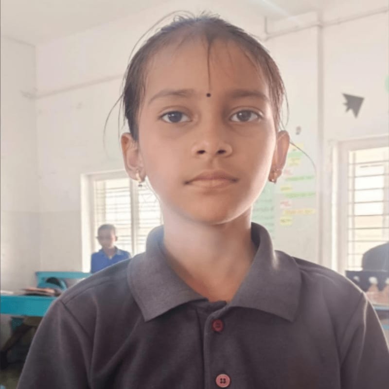 A young girl with a bindi on her forehead stands in the foreground, while a classroom with a window and other students is visible in the background.
