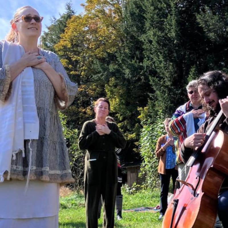 In the foreground, a woman in a striped dress and sunglasses places her hands on her chest while a man plays a cello, and in the background, several people stand with their hands on their chests in a grassy, tree-lined area.