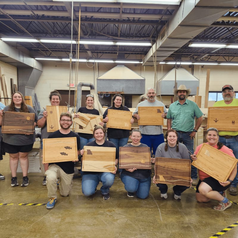 A group of people proudly display their handmade wooden cutting boards in a workshop.