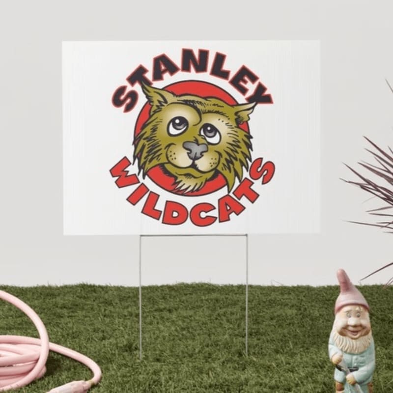 A white yard sign with a cartoon bobcat and the words "STANLEY WILDCATS" is planted in green grass, with a gnome and a pink hose in the foreground.