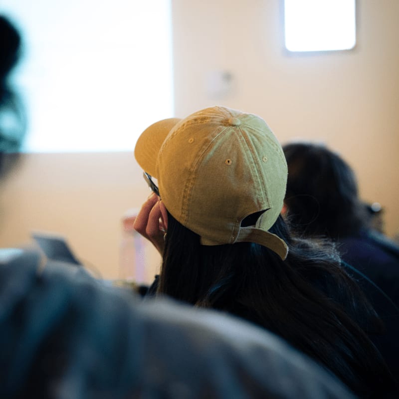 A person wearing a yellow baseball cap is in the foreground, facing away from the camera towards a bright, out-of-focus background with a projector screen.