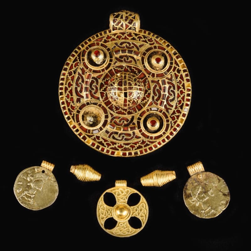 A large, ornate gold pendant with red and amber inlays sits above a collection of smaller gold artifacts, including coins and beads, all against a black background.