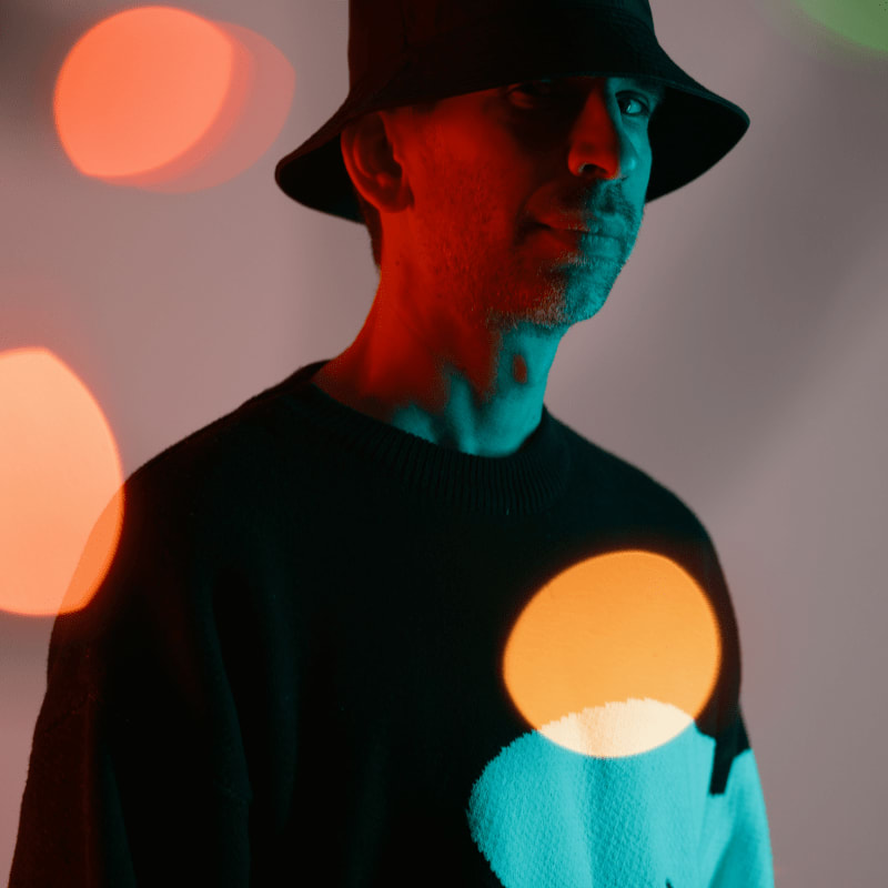 A man in a bucket hat and sweater is illuminated by colorful bokeh lights in the foreground against a plain background.