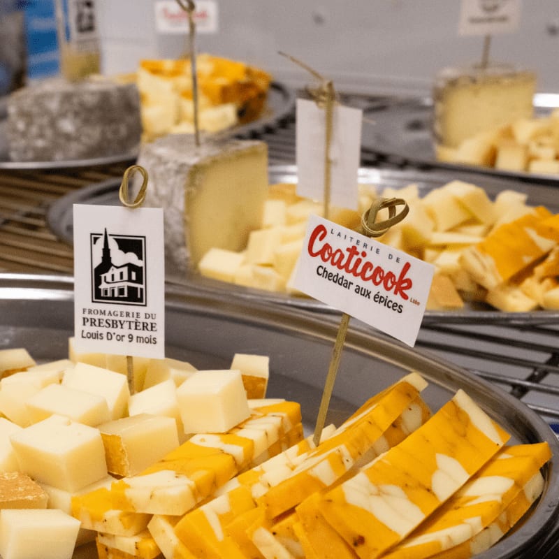 In the foreground, sliced and cubed cheeses with orange marbling are displayed on silver trays, while the background shows more cheese varieties and labels.