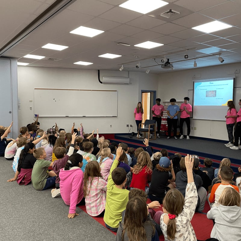 Children with raised hands sit on the floor in the foreground, while teenagers in pink shirts present on a stage in the background.