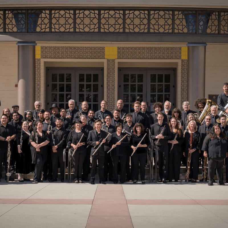 A large orchestra stands in front of a building with ornate windows, holding their instruments.