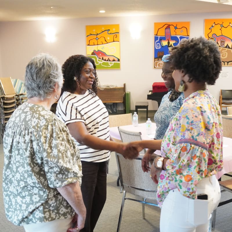 Four women are gathered in a room with colorful artwork on the walls, as two of them shake hands in the foreground.
