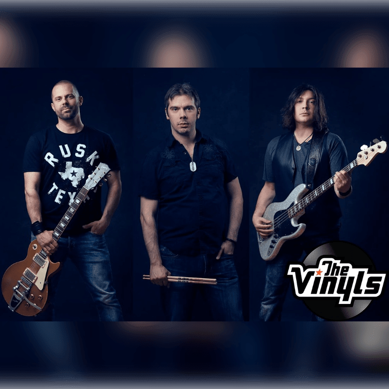 Three men, two holding guitars and one holding drumsticks, stand against a dark, blurred background with a band logo in the lower right corner.